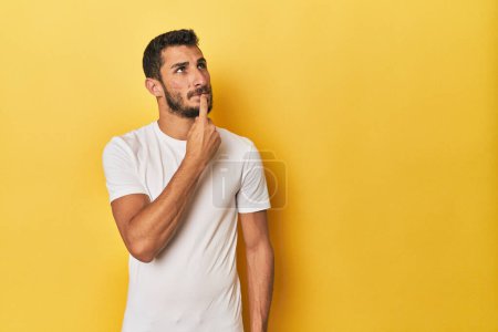 Téléchargez les photos : Jeune homme hispanique sur fond jaune regardant de côté avec une expression douteuse et sceptique. - en image libre de droit