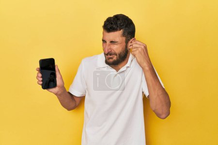 Téléchargez les photos : Jeune homme hispanique montre écran de téléphone couvrant les oreilles avec les mains. - en image libre de droit
