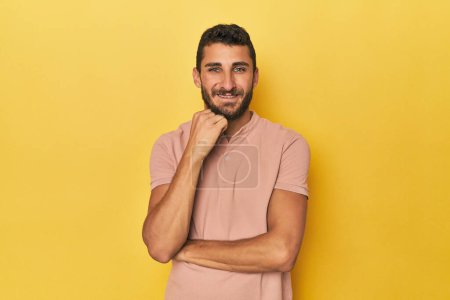 Foto de Joven hispano sobre fondo amarillo sonriendo feliz y confiado, tocando la barbilla con la mano. - Imagen libre de derechos