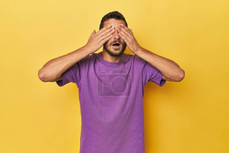 Photo for Young Hispanic man on yellow background covers eyes with hands, smiles broadly waiting for a surprise. - Royalty Free Image