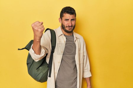 Foto de Joven hispano con mochila de viaje mostrando puño a cámara, expresión facial agresiva. - Imagen libre de derechos