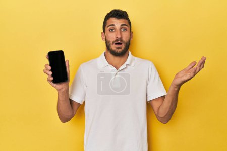 Photo for Young Hispanic man shows phone screen receiving a pleasant surprise, excited and raising hands. - Royalty Free Image