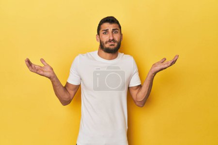 Téléchargez les photos : Jeune homme hispanique sur fond jaune doutant et haussant les épaules en questionnant geste. - en image libre de droit