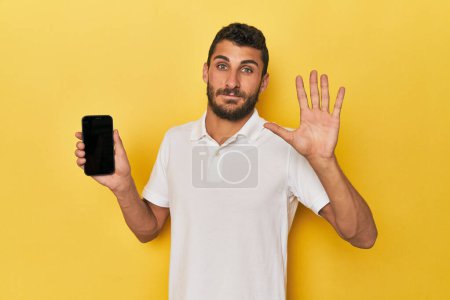 Foto de Joven hispano muestra la pantalla del teléfono sonriendo alegre mostrando el número cinco con los dedos. - Imagen libre de derechos