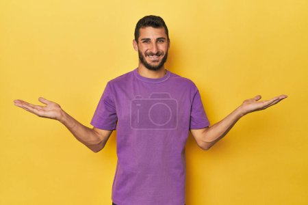 Téléchargez les photos : Jeune homme hispanique sur fond jaune fait échelle avec les bras, se sent heureux et confiant. - en image libre de droit