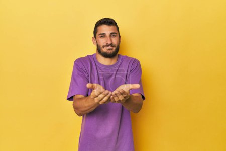 Foto de Joven hispano sobre fondo amarillo sosteniendo algo con palmas, ofreciendo a la cámara. - Imagen libre de derechos