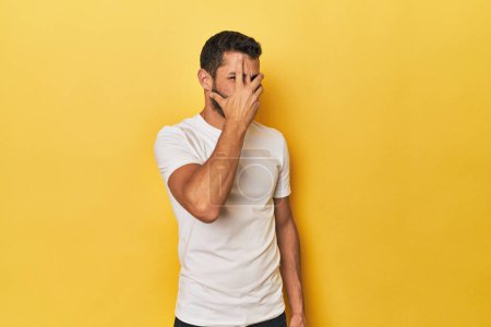 Téléchargez les photos : Jeune homme hispanique sur fond jaune cligner des yeux à la caméra par les doigts, embarrassé couvrant le visage. - en image libre de droit