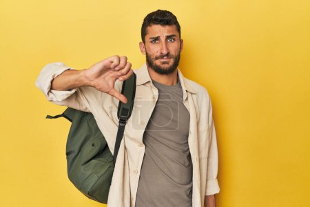 Téléchargez les photos : Jeune homme hispanique avec un sac à dos de voyage montrant un geste détestable, pouces baissés. Concept de désaccord. - en image libre de droit