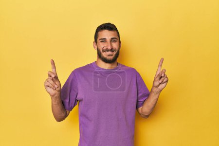 Téléchargez les photos : Jeune homme hispanique sur fond jaune indique avec les deux doigts avant montrant un espace vide. - en image libre de droit