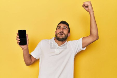 Photo for Young Hispanic man shows phone screen raising fist after a victory, winner concept. - Royalty Free Image