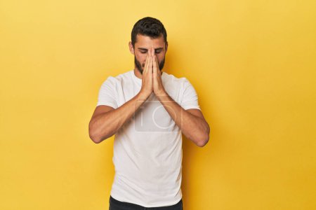 Photo for Young Hispanic man on yellow background holding hands in pray near mouth, feels confident. - Royalty Free Image