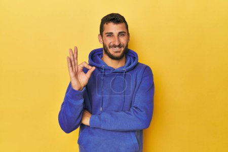 Téléchargez les photos : Jeune homme hispanique sur fond jaune clin d'oeil et tient un geste correct avec la main. - en image libre de droit