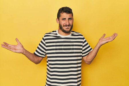 Téléchargez les photos : Jeune homme hispanique sur fond jaune épaules confuses et douteuses haussant pour tenir un espace de copie. - en image libre de droit