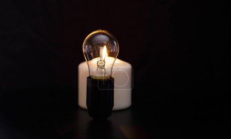 Photo for Burning candle near a switched off light bulb in dark home. Blackout, electricity off, load shedding energy crisis or power outage, concept image. - Royalty Free Image