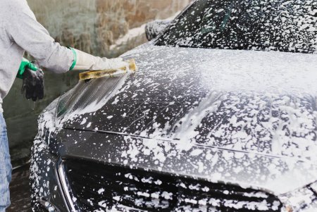 Male worker washing car at car station outdoors with foam and yellow sponge