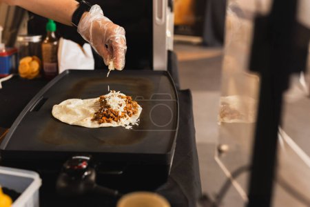Téléchargez les photos : Le chef saupoudre une tortilla de fromage préparant une quesadilla fraîche sur un gril noir - en image libre de droit