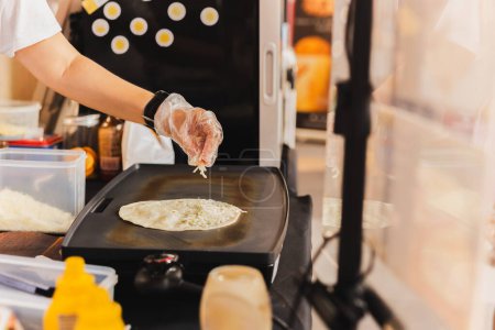 Téléchargez les photos : Le chef saupoudre une tortilla de fromage préparant une quesadilla fraîche sur un gril noir - en image libre de droit