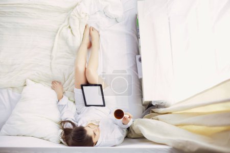 Téléchargez les photos : Portrait de femme utilisant un comprimé et buvant du café assis sur le lit le matin. - en image libre de droit