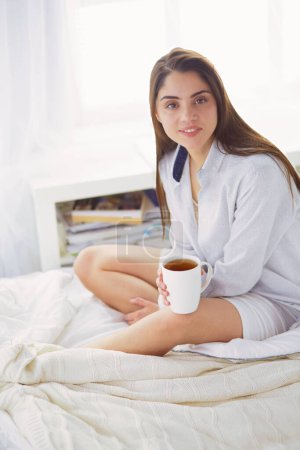 Téléchargez les photos : Femme assise au lit lisant un livre et prenant le petit déjeuner. - en image libre de droit