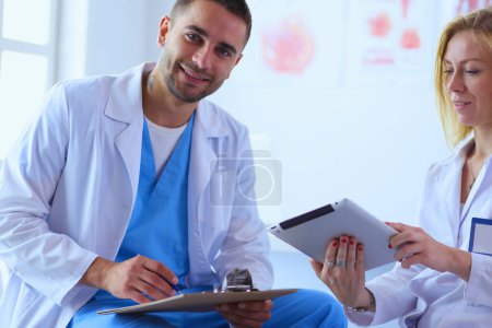 Foto de El guapo doctor está hablando con una joven doctora y tomando notas mientras está sentado en su consultorio. - Imagen libre de derechos