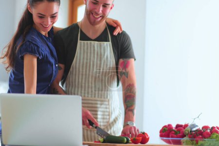 Téléchargez les photos : Jeune homme coupant des légumes et femme debout avec ordinateur portable dans la cuisine. - en image libre de droit