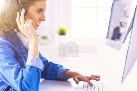 Téléchargez les photos : Heureuse charmante jeune femme assise et travaillant avec un ordinateur portable en utilisant un casque dans le bureau. - en image libre de droit