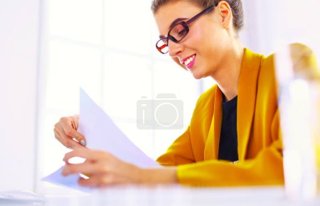 Foto de Hermosa joven mujer de negocios haciendo un poco de papeleo mientras está sentado en el escritorio de la oficina delante de la computadora portátil - Imagen libre de derechos