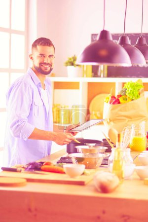 Téléchargez les photos : Homme suivant la recette sur tablette numérique et la cuisine savoureuse et saine dans la cuisine à la maison. - en image libre de droit