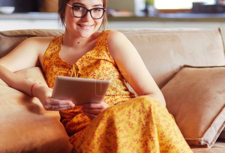 Foto de Hermosa joven morena en casa sentada en un sofá o sofá usando su tablet y sonriendo
. - Imagen libre de derechos