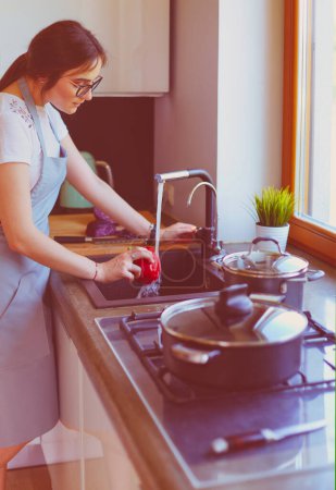 Téléchargez les photos : Femme lavant les légumes. Belle jeune femme lavant les légumes pour salade et souriant tout en se tenant dans la cuisine. - en image libre de droit