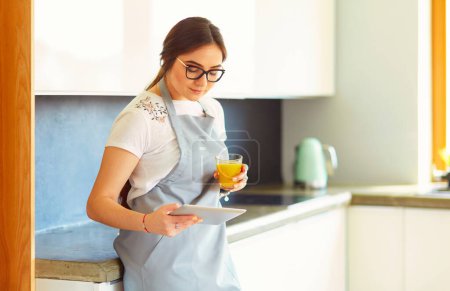Téléchargez les photos : Jeune femme avec jus d'orange et tablette dans la cuisine. - en image libre de droit