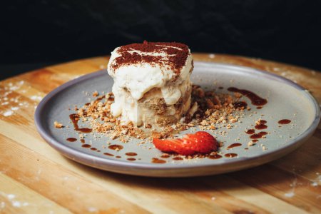 Tiramisu dessert topped with cocoa powder alongside crushed biscuits and strawberry garnish on a wooden table.