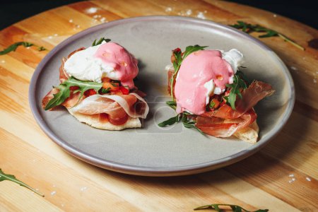 Two perfectly poached eggs sit atop golden english muffins, drizzled with hollandaise sauce and garnished with a dollop of pink sour cream on a white plate.