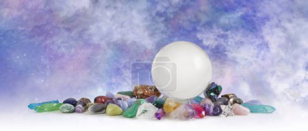 Large clear crystal ball celestial crystal healing banner - skrying sphere sitting atop selection of tumbled healing stones in front of heavenly sky background with copy space 