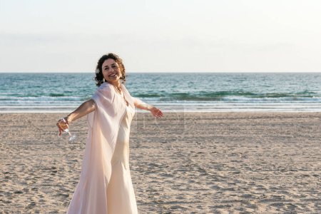 Eine Frau in Weiß mit offenen Armen an einem ruhigen Strand mit Blick auf den Ozean.