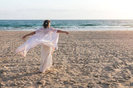 Eine Person in Weiß mit offenen Armen an einem ruhigen Strand mit Blick auf den Ozean.