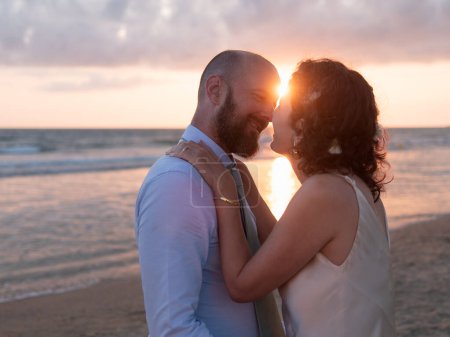 A couple embraces on a beach, the sun setting behind them casts a warm glow.