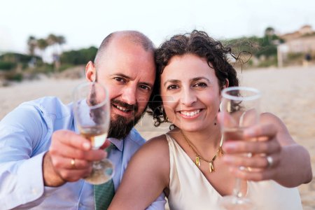 A couple toasts with wine glasses on a beach at sunset, celebrating a special moment.