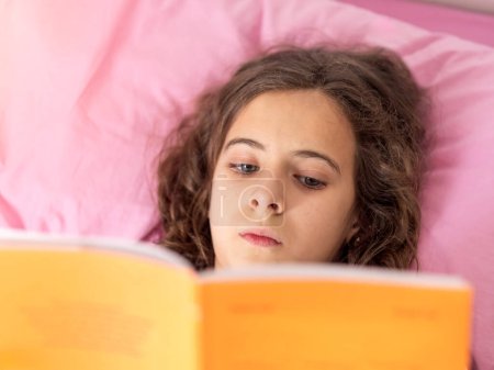 Une jeune fille absorbée dans un livre tout en étant confortablement couchée dans un lit rose.