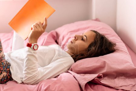Téléchargez les photos : Une jeune fille absorbée dans un livre tout en étant confortablement couchée dans un lit rose. - en image libre de droit