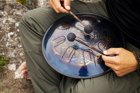 Photo for Caucasian musician hands holding drum sticks and playing a modern hand pan steel tongue drum percussion instrument on nature background - Royalty Free Image