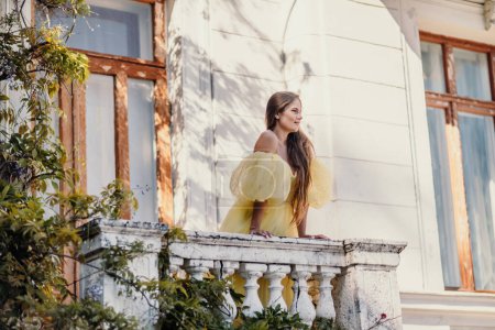 Téléchargez les photos : Une belle femme souriante et gentille dans une magnifique robe jaune se tient sur le balcon d'une vieille maison vintage. - en image libre de droit