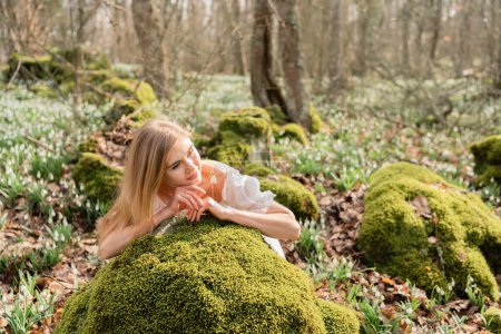 Photo for Snowdrops galanthus blond. A girl in a white dress lay down on a stone in the moss in a meadow with snowdrops in a spring forest. - Royalty Free Image