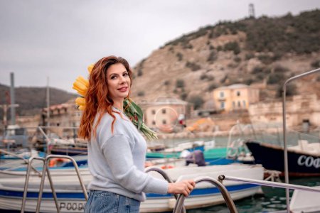 Woman holds yellow tulips in harbor with boats docked in the background., overcast day, yellow sweater, mountains.
