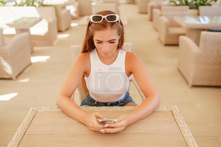 Woman sits in outdoor cafe at wooden table, holds smartphone. Bright sunny day provides natural lighting. Cafe offers relaxed atmosphere for customers to enjoy refreshments