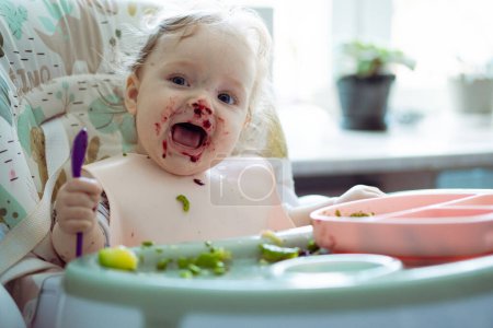 Téléchargez les photos : Bébé fille mignonne essaie de manger son premier repas. Le sevrage dirigé par bébé (BLW). Adorable bébé mangeant dans la chaise bébé - en image libre de droit
