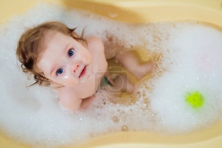 Téléchargez les photos : Bébé fille dans un bain avec mousse et bulles de savon. Joyeux bébé riant prenant un bain jouant avec des jouets. Petit enfant dans une baignoire. Enfant souriant dans la salle de bain. Lavage et baignade des nourrissons - en image libre de droit