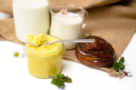 Ghee butter in a jar, milk and swirl bun at sackcloth. Clarified butter and bun on white background. Organic, healthy food. Cooking oil, pure ghee. Healthy fat diet concept.