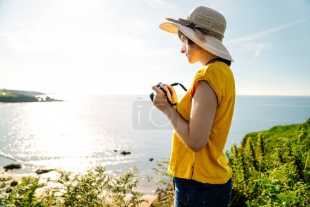 Photo for A casually dressed blonde tourist in a vibrant yellow t-shirt and hat, capturing a scenic coastal landscape from a lush green cliff with noonday light - Royalty Free Image