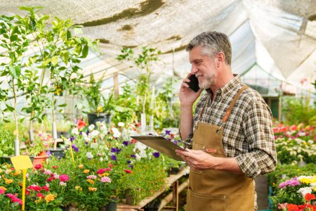 Photo for A close-up photo of a floral nursery worker speaking on a mobile phone, holding a folder while attending to a customer's flower order. - Royalty Free Image